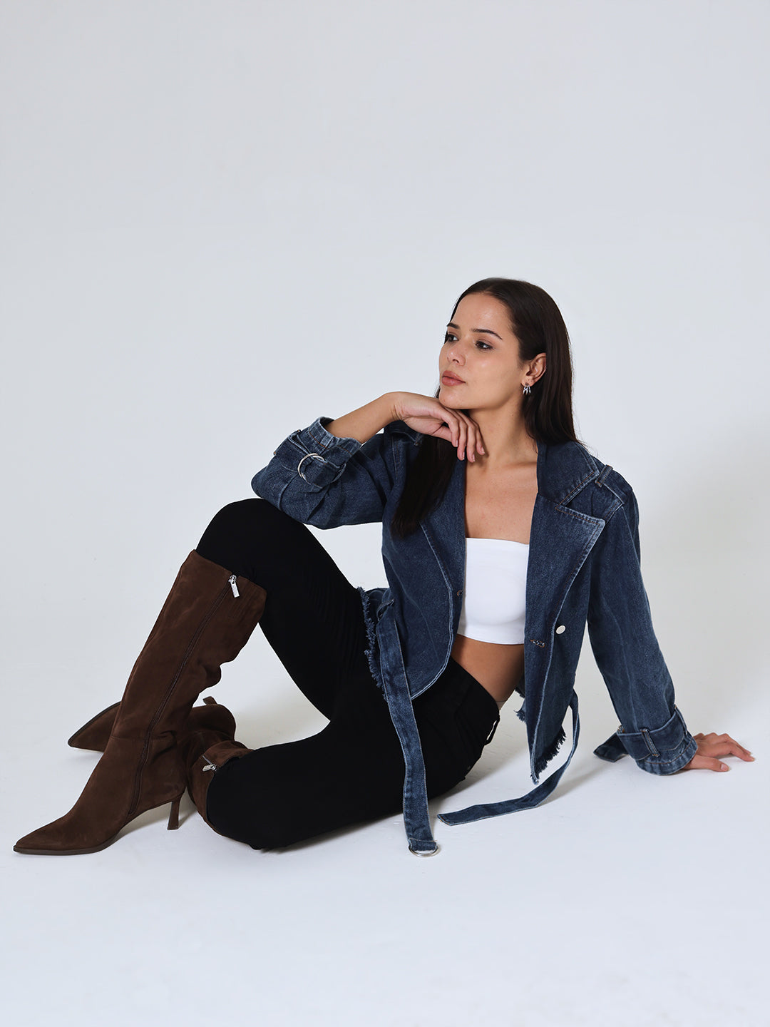 Woman sitting on a white floor wearing Dvrzi's denim biker jacket, black pants, and brown knee-high boots.